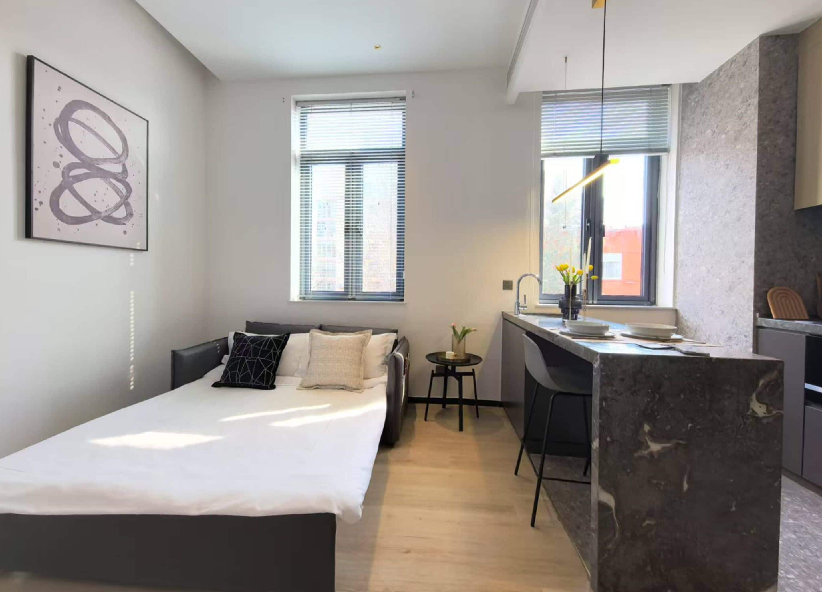 Ein modernes Schlafzimmer mit Bett, Schreibtisch und einem kleinen Hocker. Ein Fenster mit Jalousien und ein Bild an der Wand.