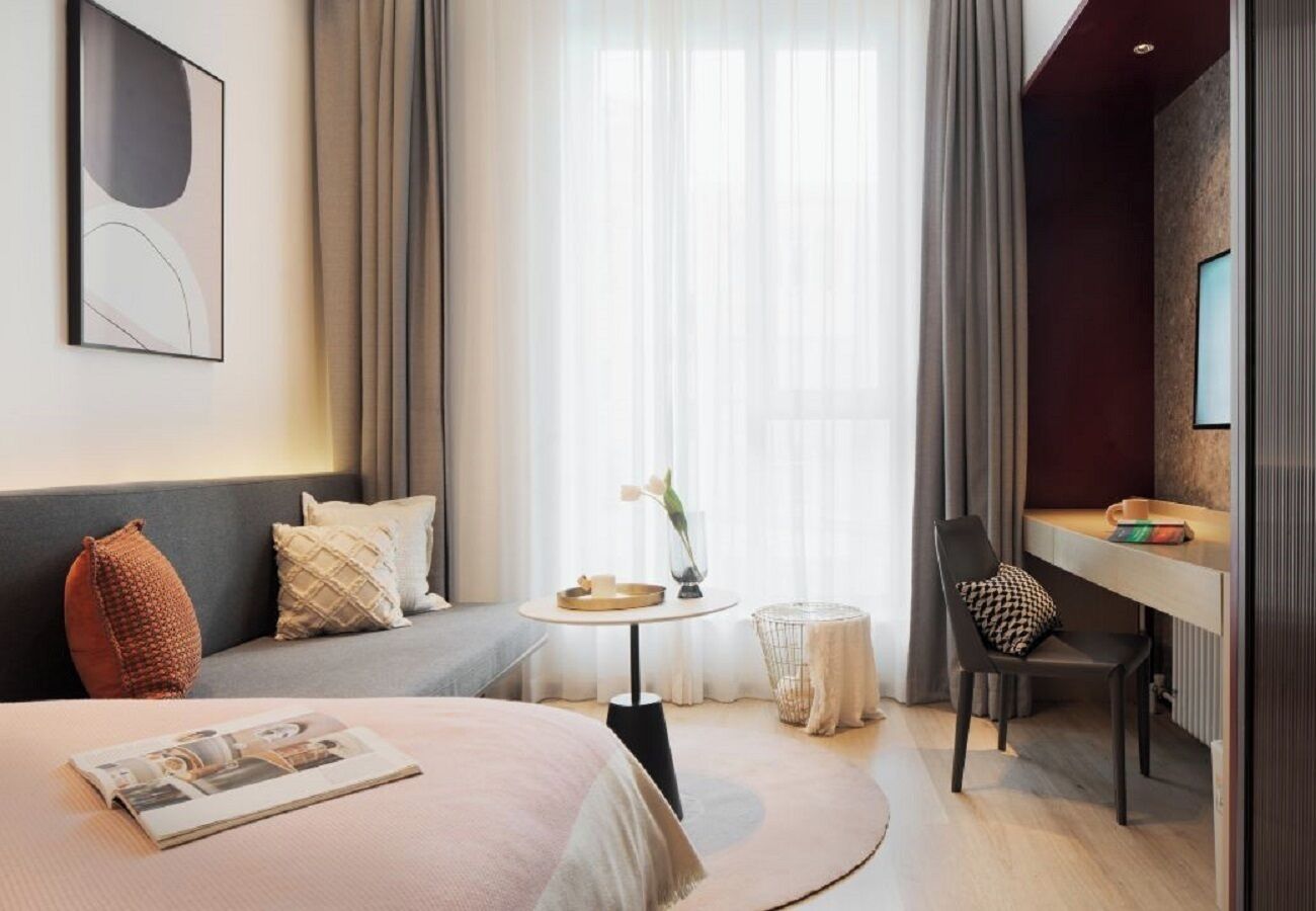 A hotel room with a bed, a desk, a chair, a small table with a vase, and a window with curtains.