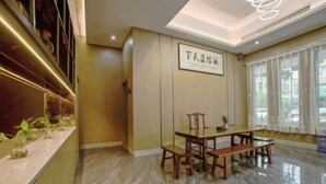 Un hall d’hôtel avec un comptoir d’accueil en bois, un espace repas comprenant une table et des bancs, et une grande fenêtre ornée de rideaux légers.