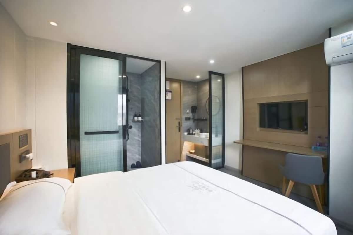 A modern hotel room with a large bed, a desk, a shower area, and a small kitchenette.