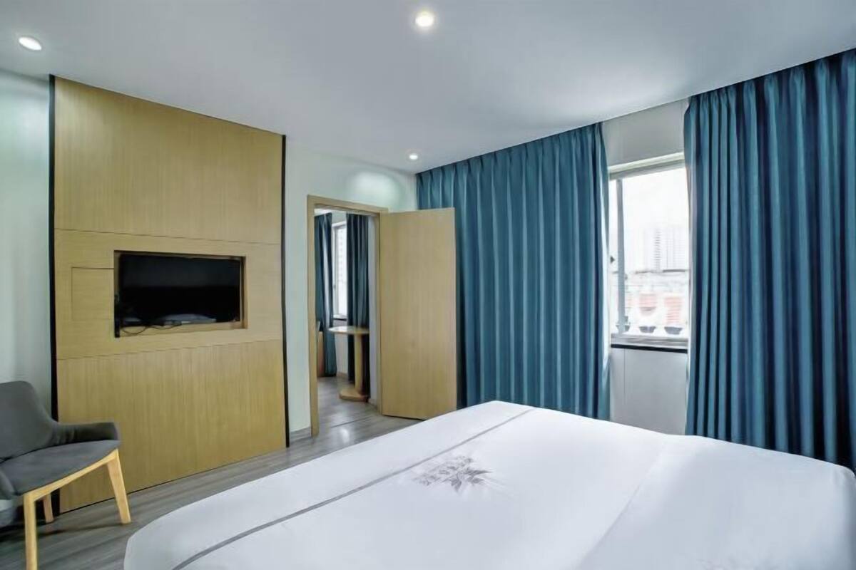 A hotel room with a bed, a TV mounted on the wall, a chair, and a view of the city through the window.