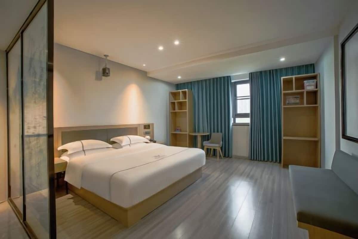A hotel room with a large bed, a desk, and a chair. There are shelves and a window with curtains.
