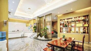 A hotel lobby with a reception desk, a staircase, a dining area with a table and chairs, and a display of items on shelves.