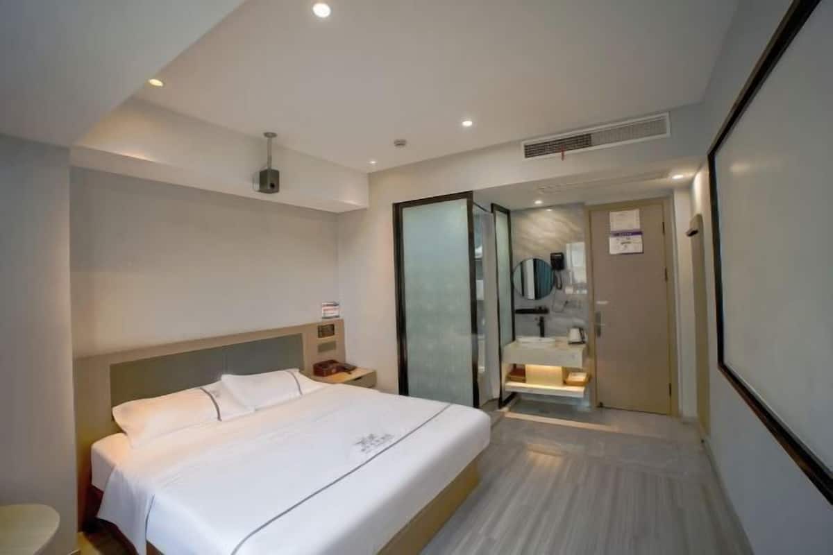 A modern hotel room with a large bed, a bedside table, a nightstand, a mirror, and a bathroom visible through a glass door.