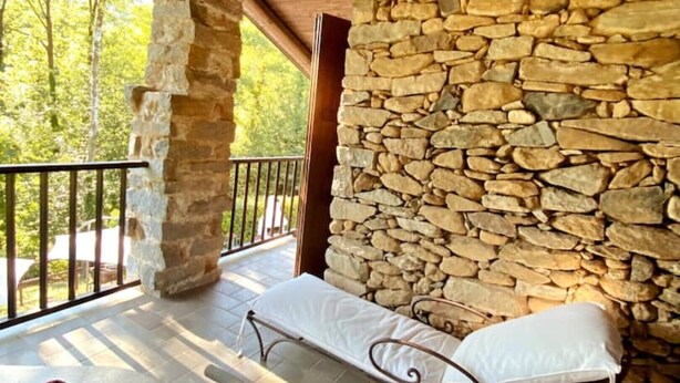 A stone wall, a wooden balcony, and a metal bench with white cushions.