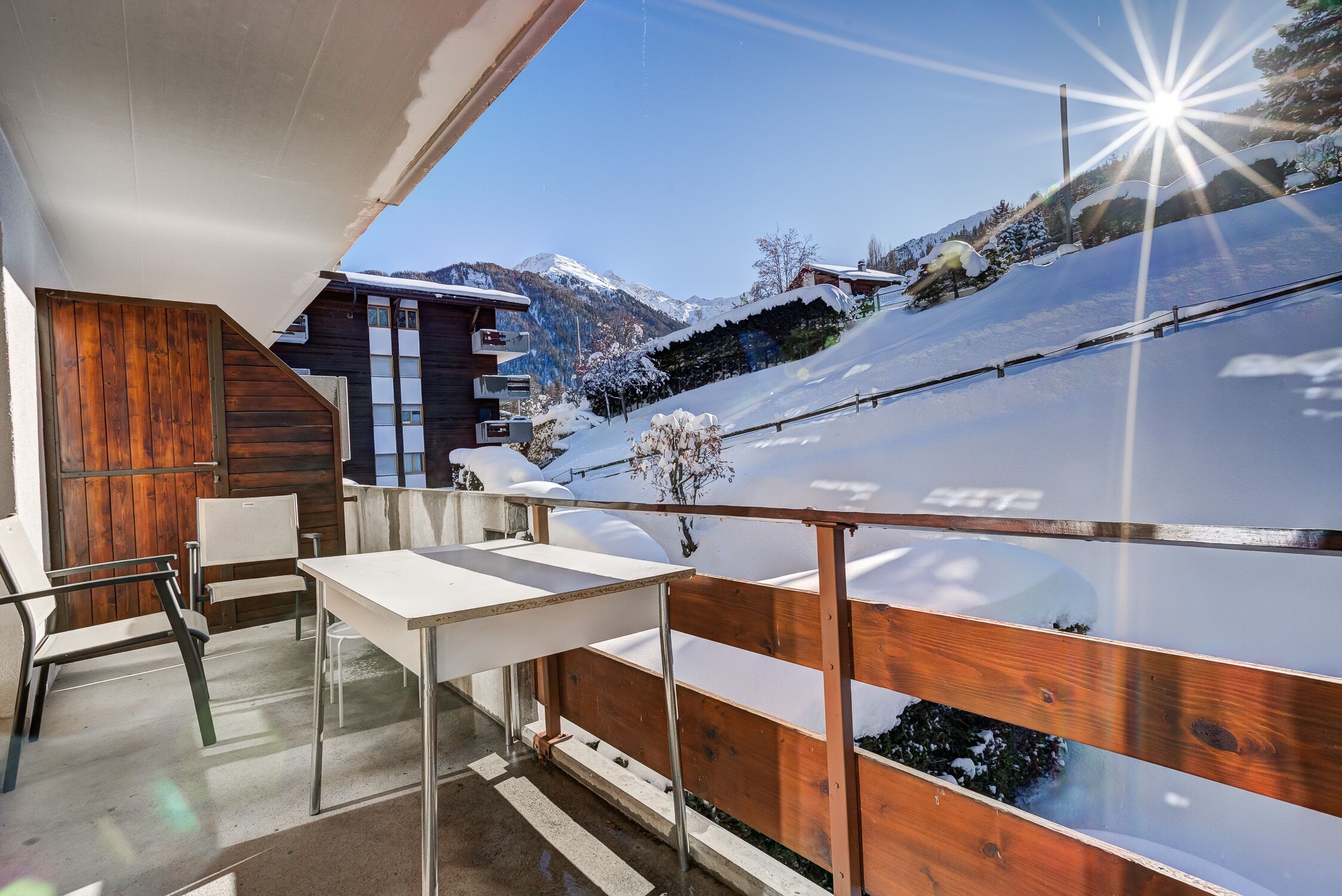 Varanda moderna ao ar livre com guarda-corpo de madeira, mesa e cadeiras, com vista para uma paisagem montanhosa coberta de neve.