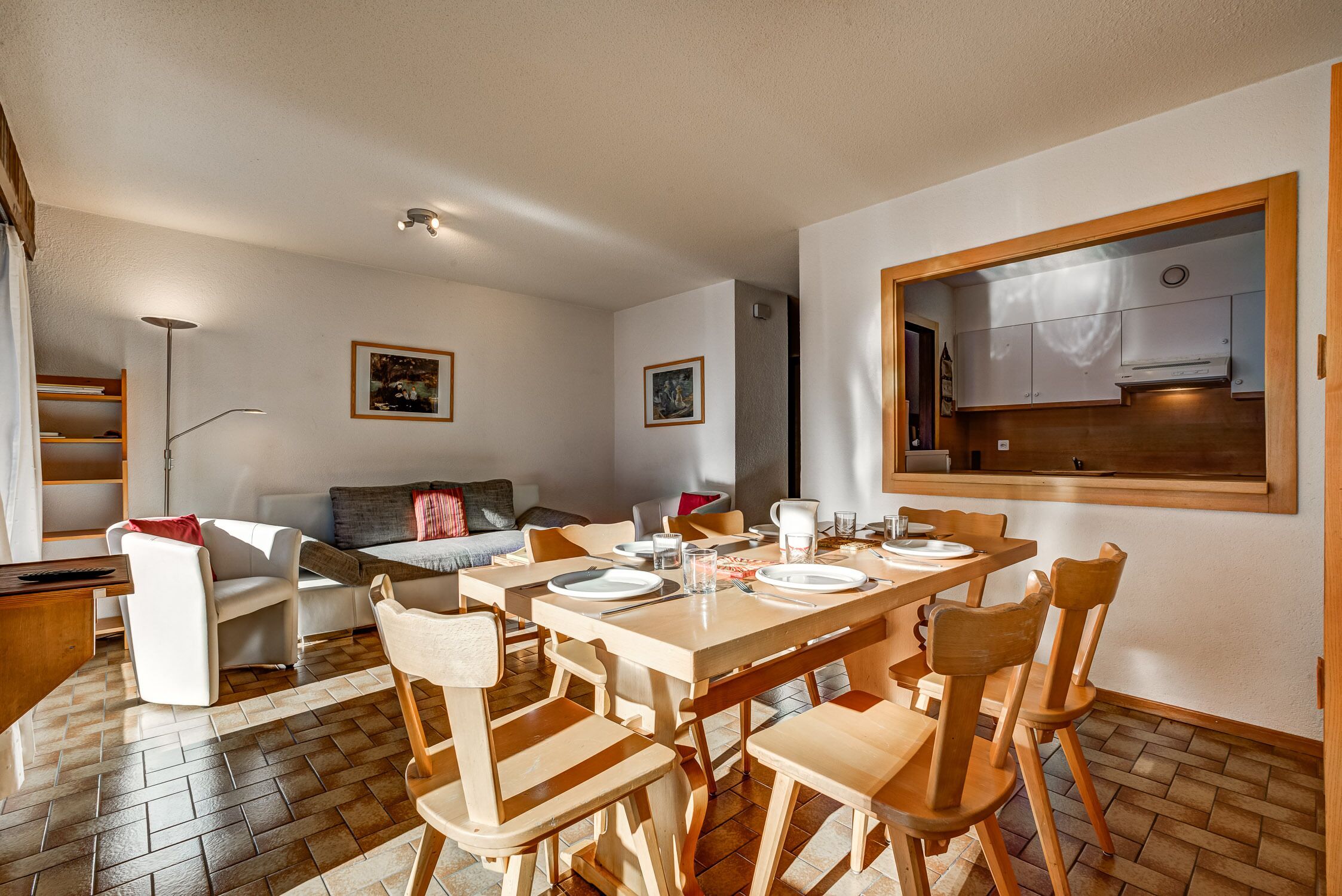 A dining area with a wooden table set for four, surrounded by wooden chairs. There is a sofa and a small table with a lamp in the background.