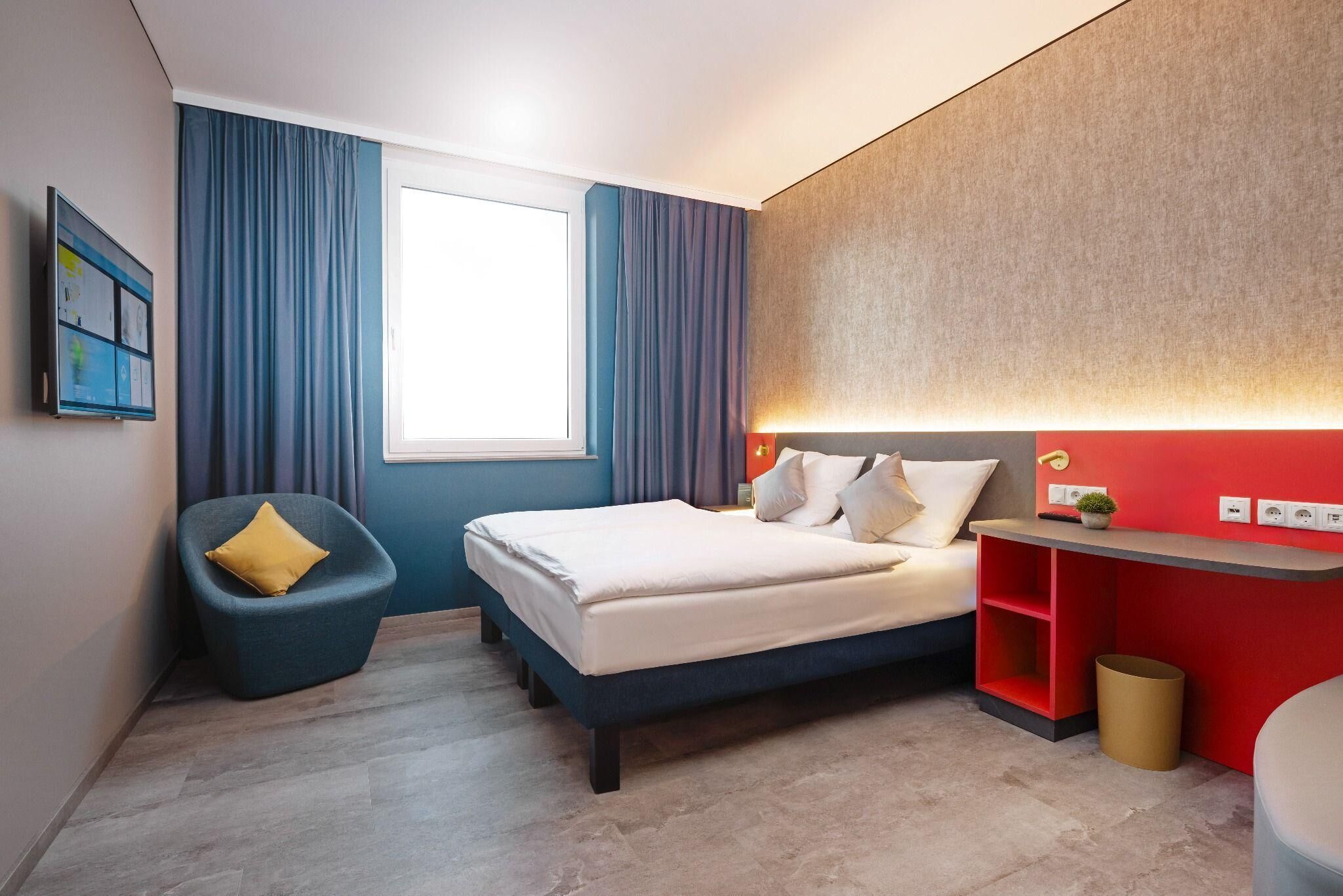A modern hotel room with a bed, a chair, a desk, and a window.