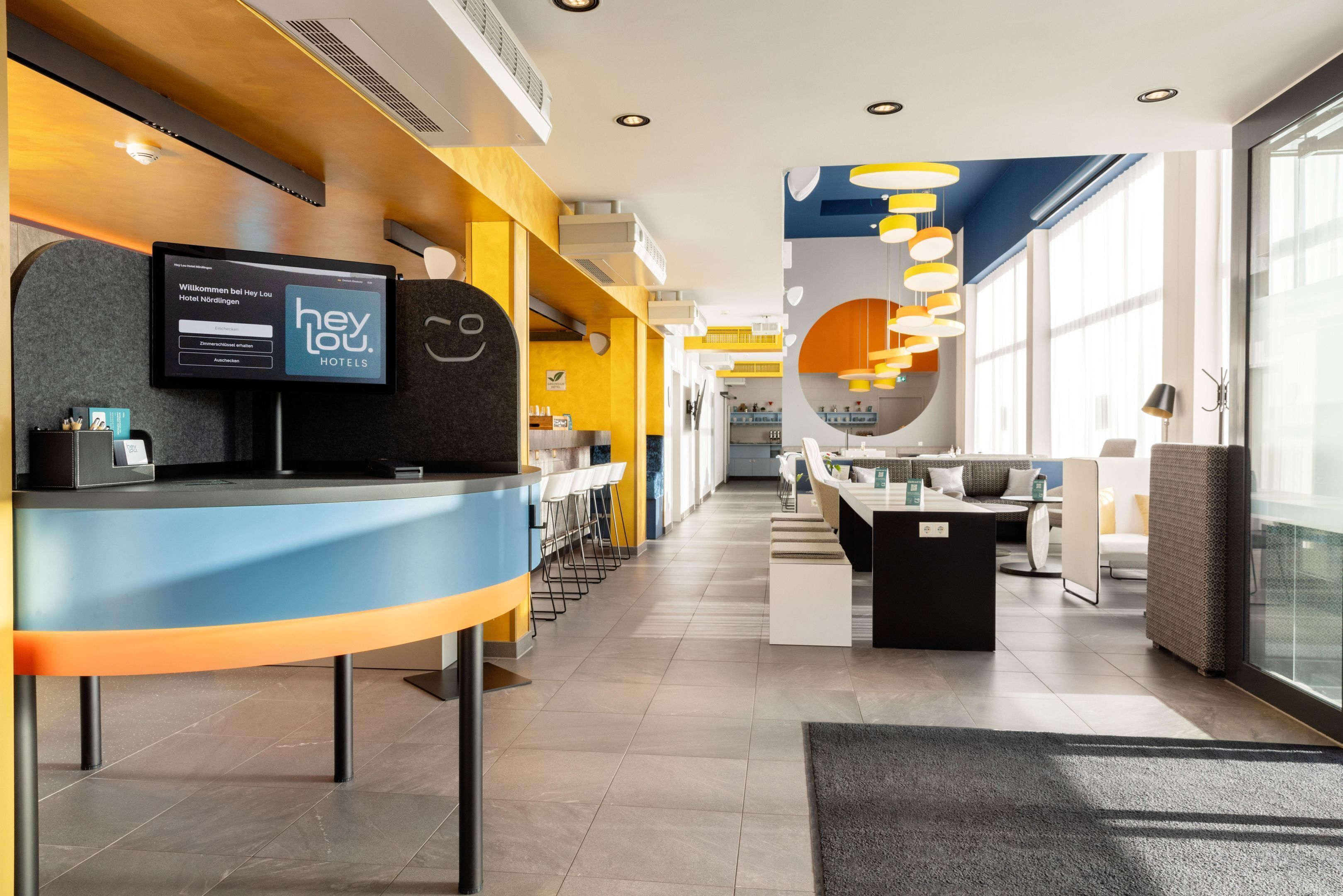 Un área de recepción de hotel moderna con una pantalla digital, un mostrador de recepción y un espacio de espera con mesas y sillas.