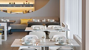 Un comedor moderno con varias mesas dispuestas para el desayuno, una cocina al fondo y un amplio ventanal con cortinas.