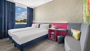 Habitación de hotel con dos camas, un escritorio, un sofá y un amplio ventanal con cortinas.