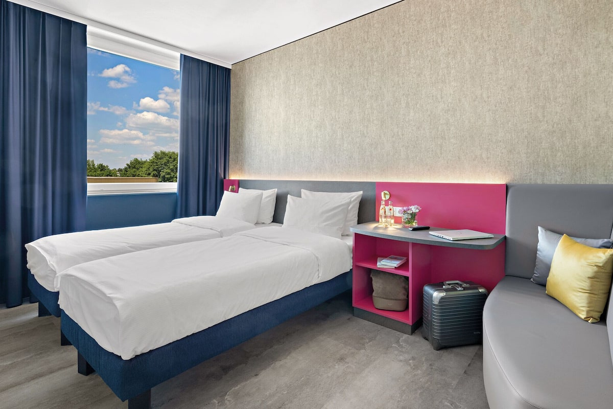 Een hotelkamer met twee bedden, een bureau, een bank en een groot raam met gordijnen.
