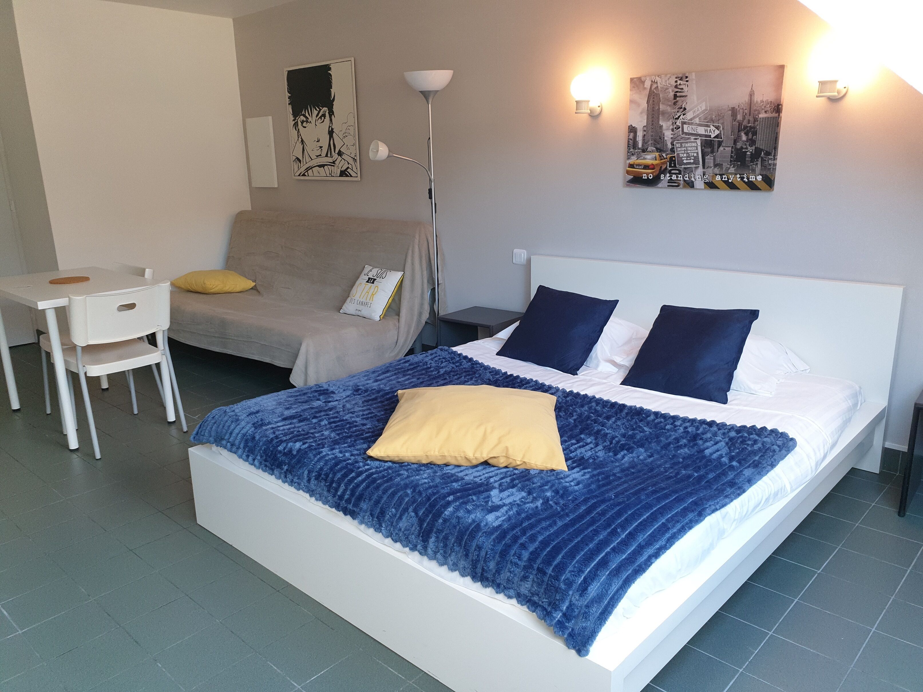 A hotel room with a bed, a sofa, a small table, and a chair. There are two wall-mounted lights and two wall art pieces.