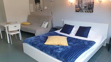 A hotel room with a bed, a sofa, a small table, and a chair. There are two wall-mounted lights and two wall art pieces.