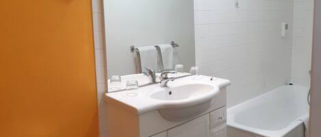 Baño con lavamanos blanco, espejo y bañera. Incluye inodoro y un cesto de basura.