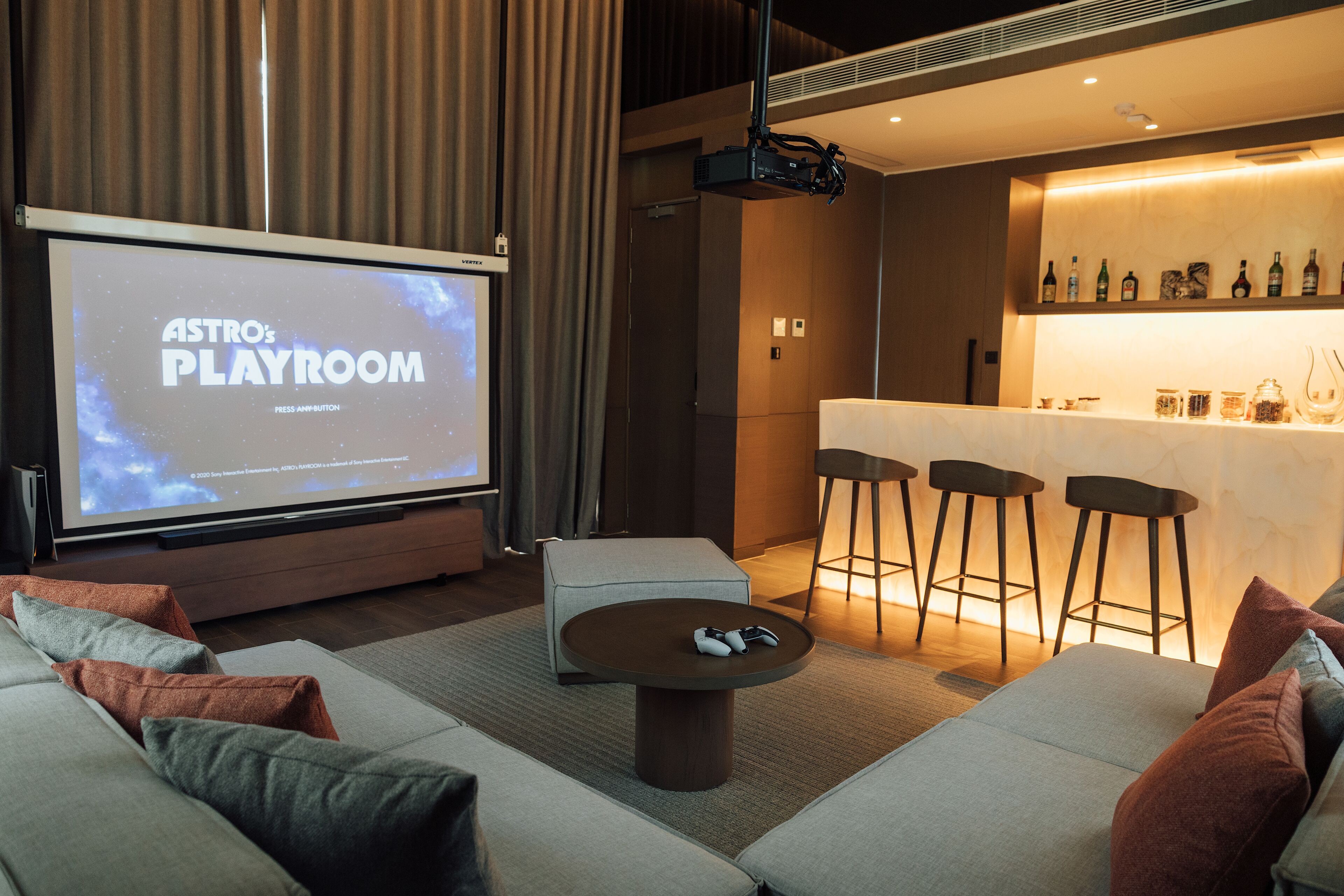 A modern living room with a large screen displaying "ASTRO'S PLAYROOM," a bar area with stools, and a sofa with cushions.
