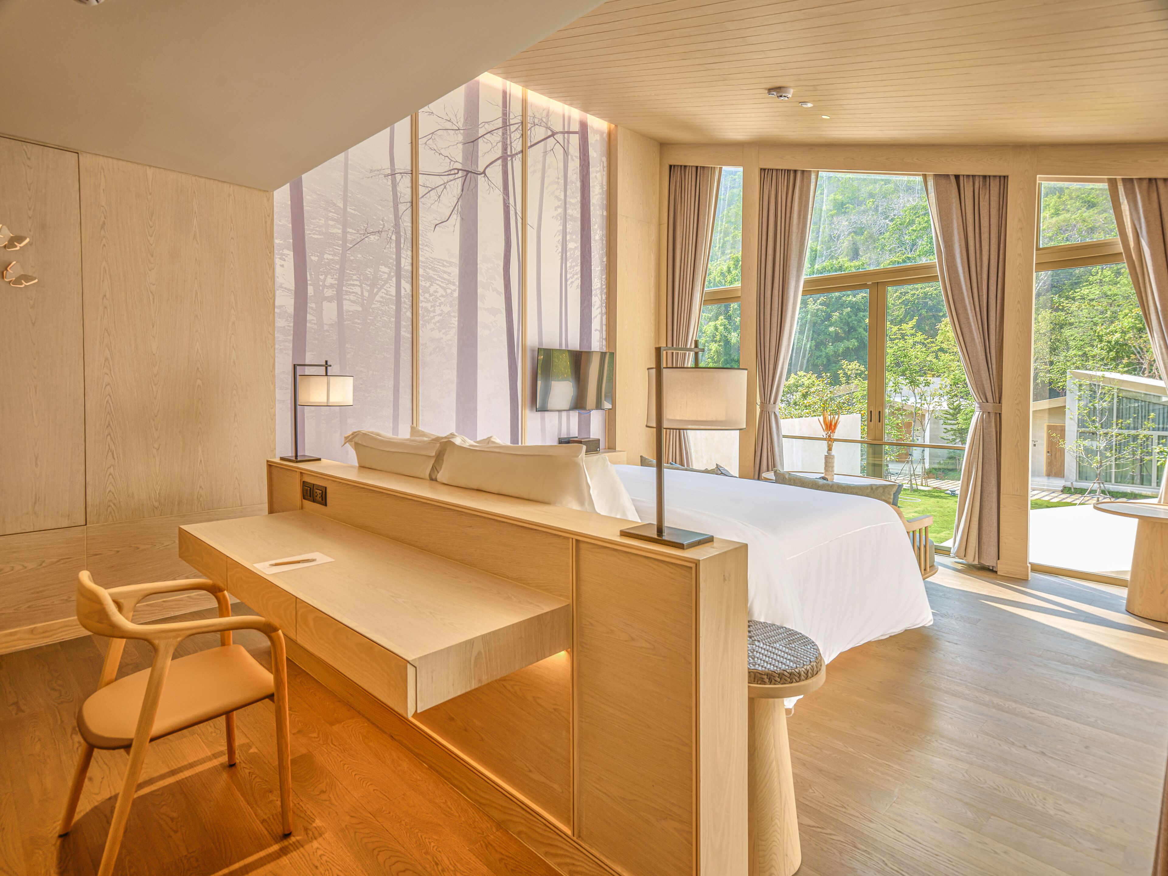 A modern bedroom with a large bed, a desk, and a chair. There are multiple windows providing natural light and views of the outdoors.