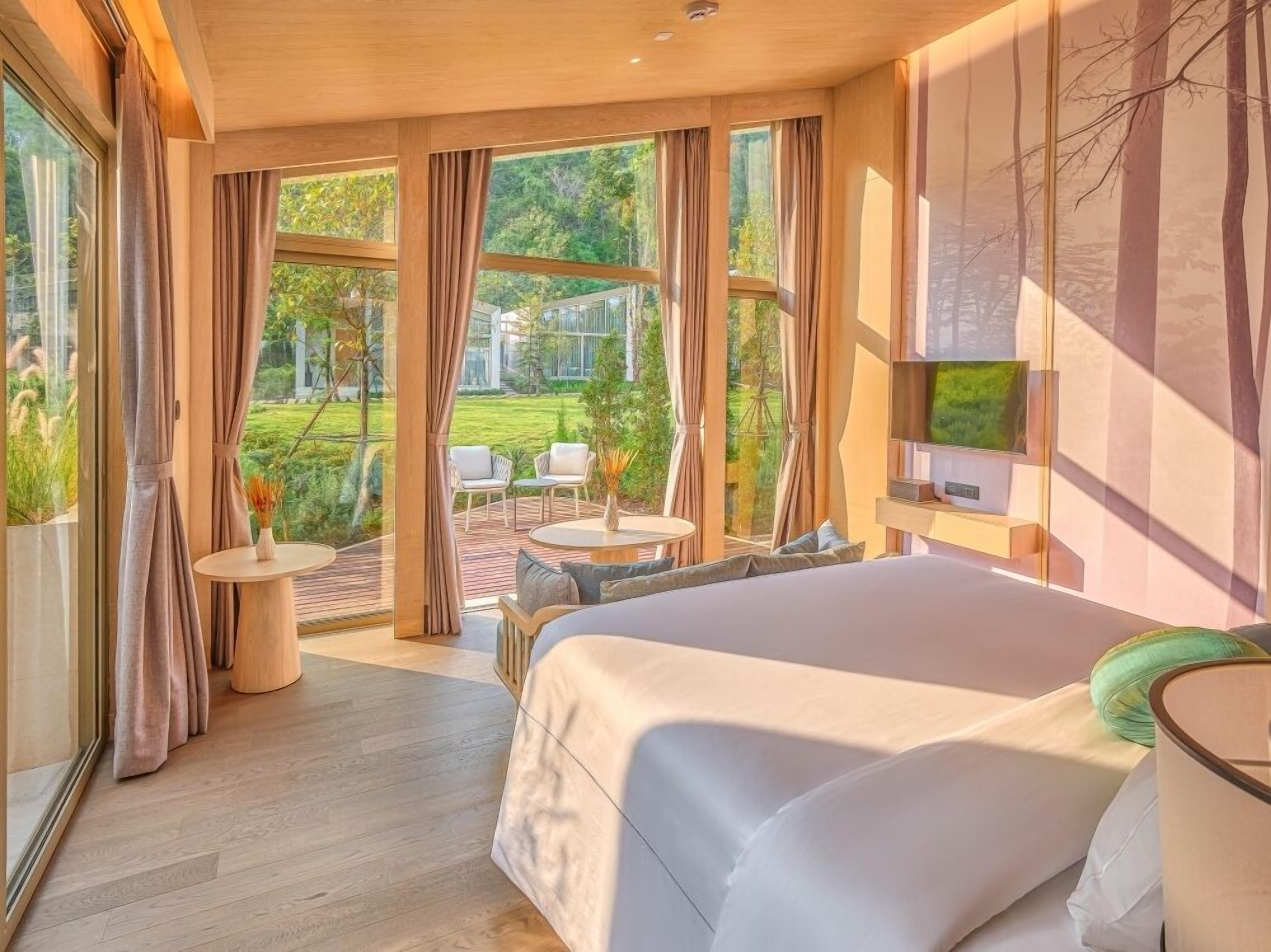 A modern hotel room with a large bed, a TV mounted on the wall, and a view of a garden through large windows.