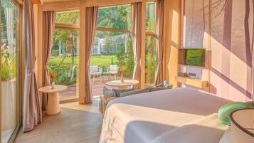 A modern hotel room with a large bed, a TV mounted on the wall, and a view of a garden through large windows.