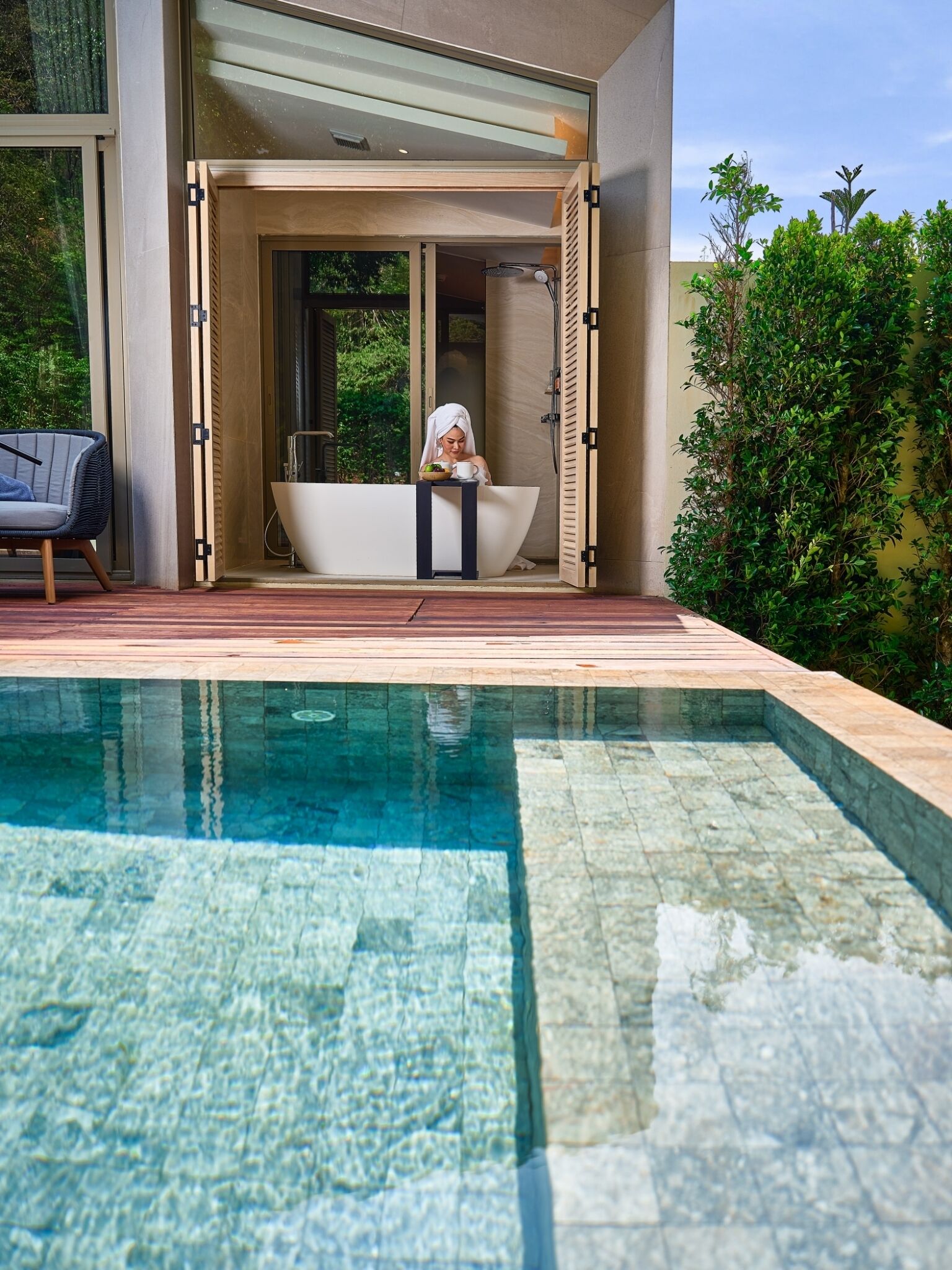 Un área de piscina moderna al aire libre con una bañera independiente, una tumbona y un área de ducha con paredes de vidrio.
