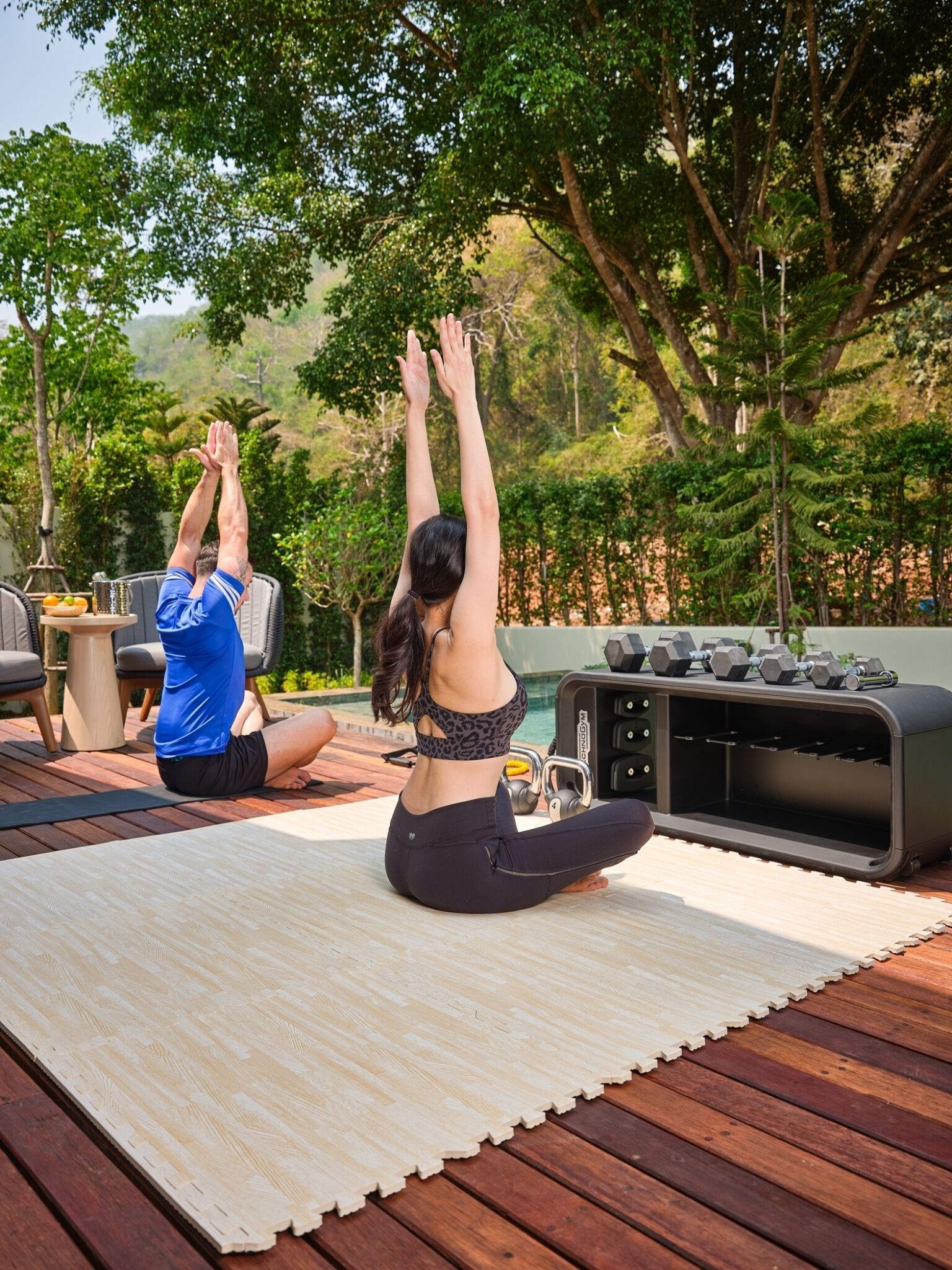 Two people performing yoga on a wooden deck with a pool and outdoor gym equipment in the background.