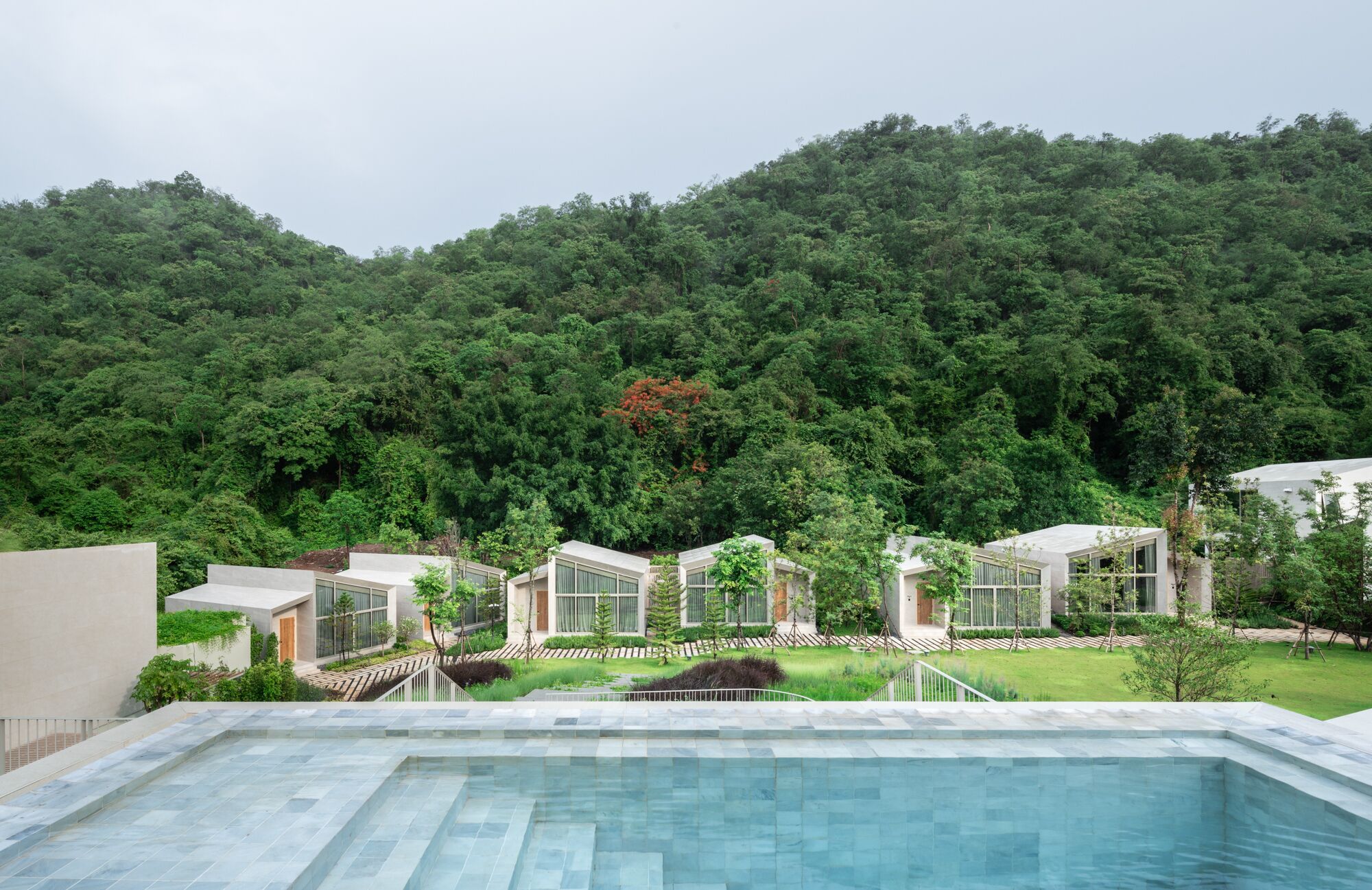A modern hotel with a pool, surrounded by lush greenery and a forested hill.