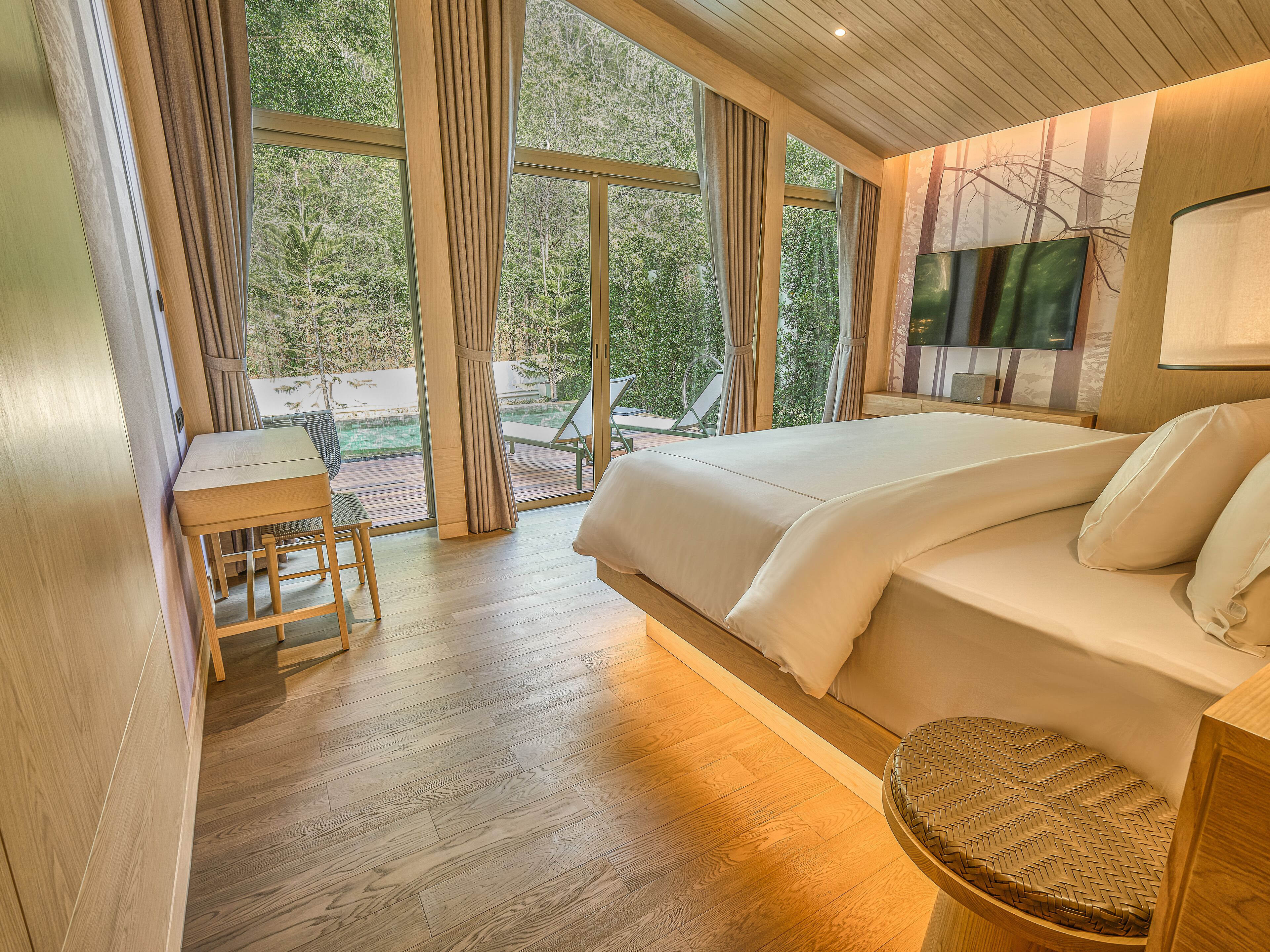 A bedroom with a large bed, a desk, and a view of the outdoors through large windows.