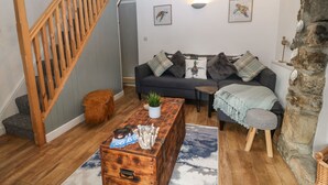 Ein gemütliches Wohnzimmer mit einer Holztreppe, einem dunkelgrauen Sofa mit gemusterten Kissen, einem rustikalen Holztisch und einer Steinwand.