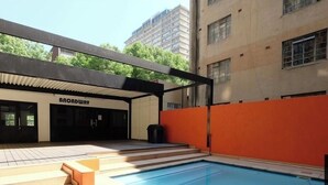An outdoor swimming pool with steps leading down to the water, surrounded by a building with a 'Broadway' sign.