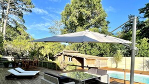 Una terraza de madera con una mesa de vidrio y sillas de mimbre blanco, una piscina y una estructura de madera al fondo.