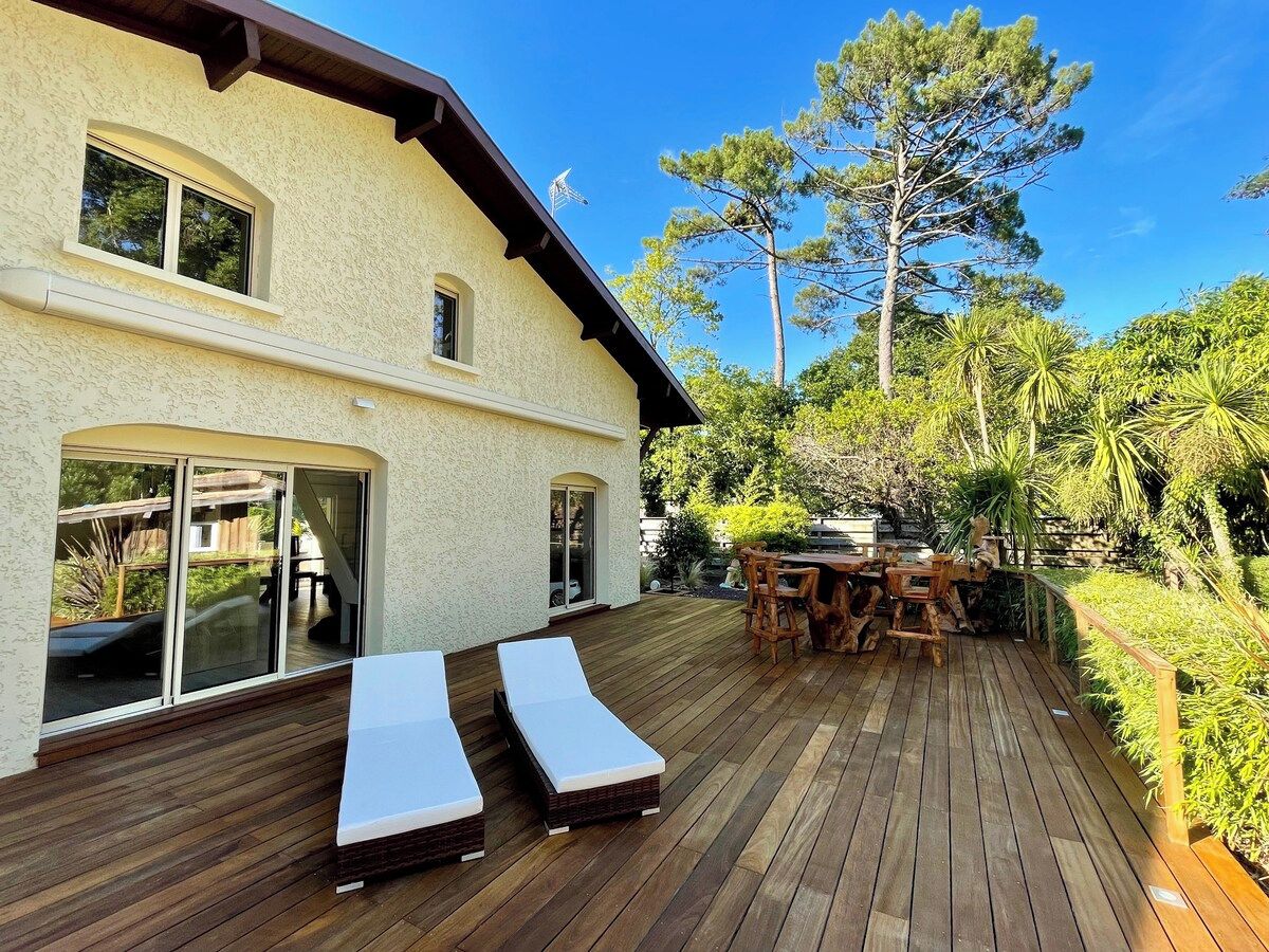 A wooden deck with lounge chairs and a dining table, surrounded by greenery and a building with large windows.