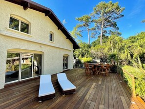 A wooden deck with lounge chairs and a dining table, surrounded by greenery and a building with large windows. - Comfortable house with heated pool near bay A0042 (Lège-Cap-Ferret)
