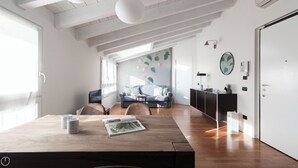 A modern living room with a wooden dining table, a sofa, and a kitchen area with a black cabinet and a white door. - Italianway Comfort - Rubens 19 B (Milan)