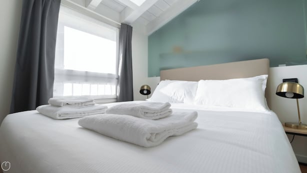A neatly made bed with white linens and folded towels, a bedside lamp, and a view of the outdoors through a window with curtains. - Italianway Comfort - Rubens 19 B (Milan)