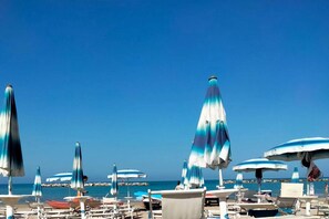 A beach with rows of sun loungers and umbrellas, a rocky shoreline, and clear blue skies.