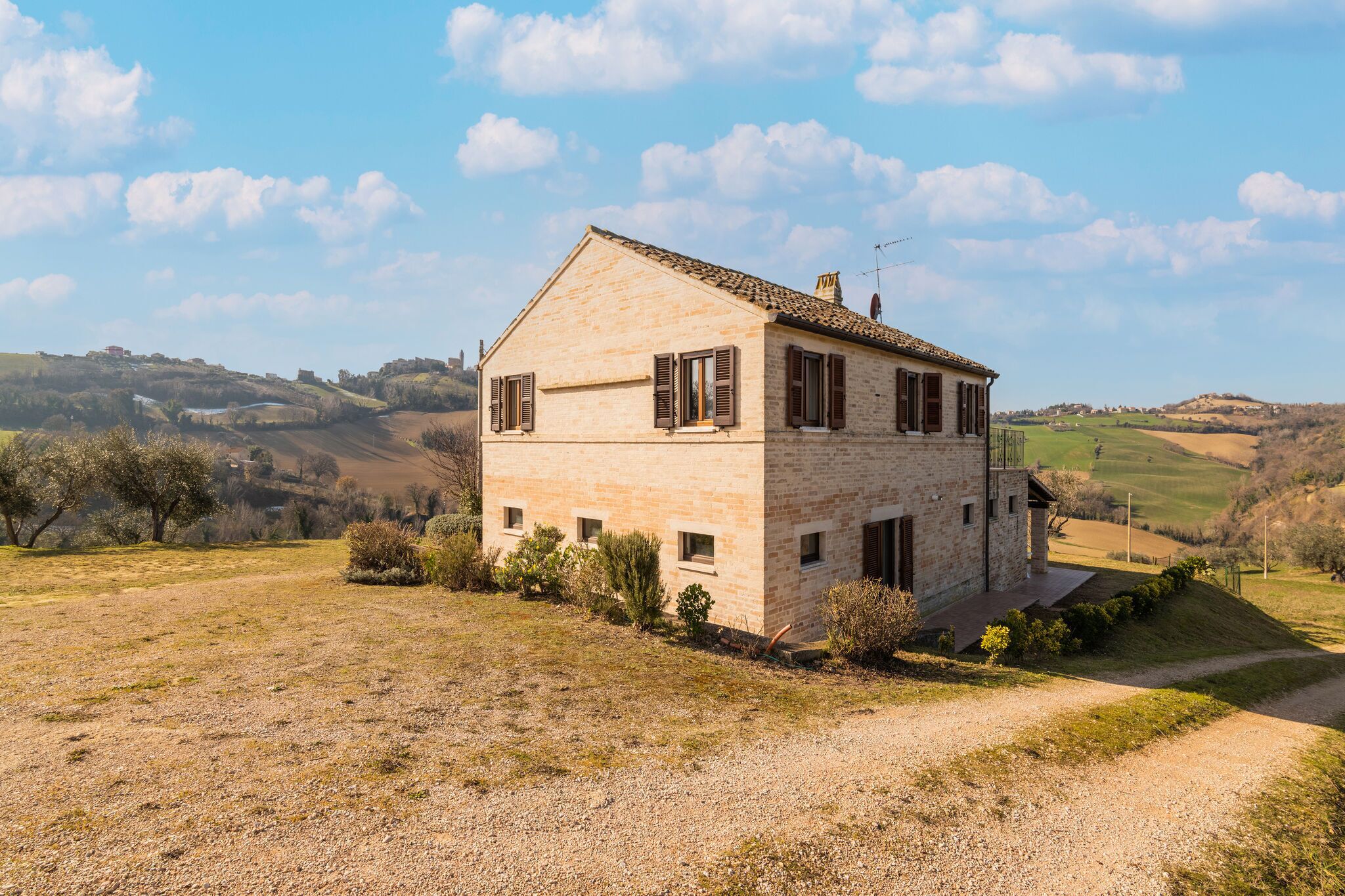 A stone house with a sloped roof, surrounded by a gravel driveway and sparse vegetation.