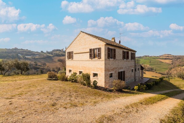 A stone house with a sloped roof, surrounded by a gravel driveway and sparse vegetation.