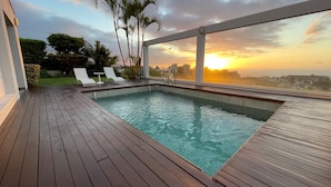 Une piscine extérieure dotée d’une terrasse en bois, de deux chaises longues et offrant une vue sur le coucher de soleil au-dessus d’un paysage urbain.