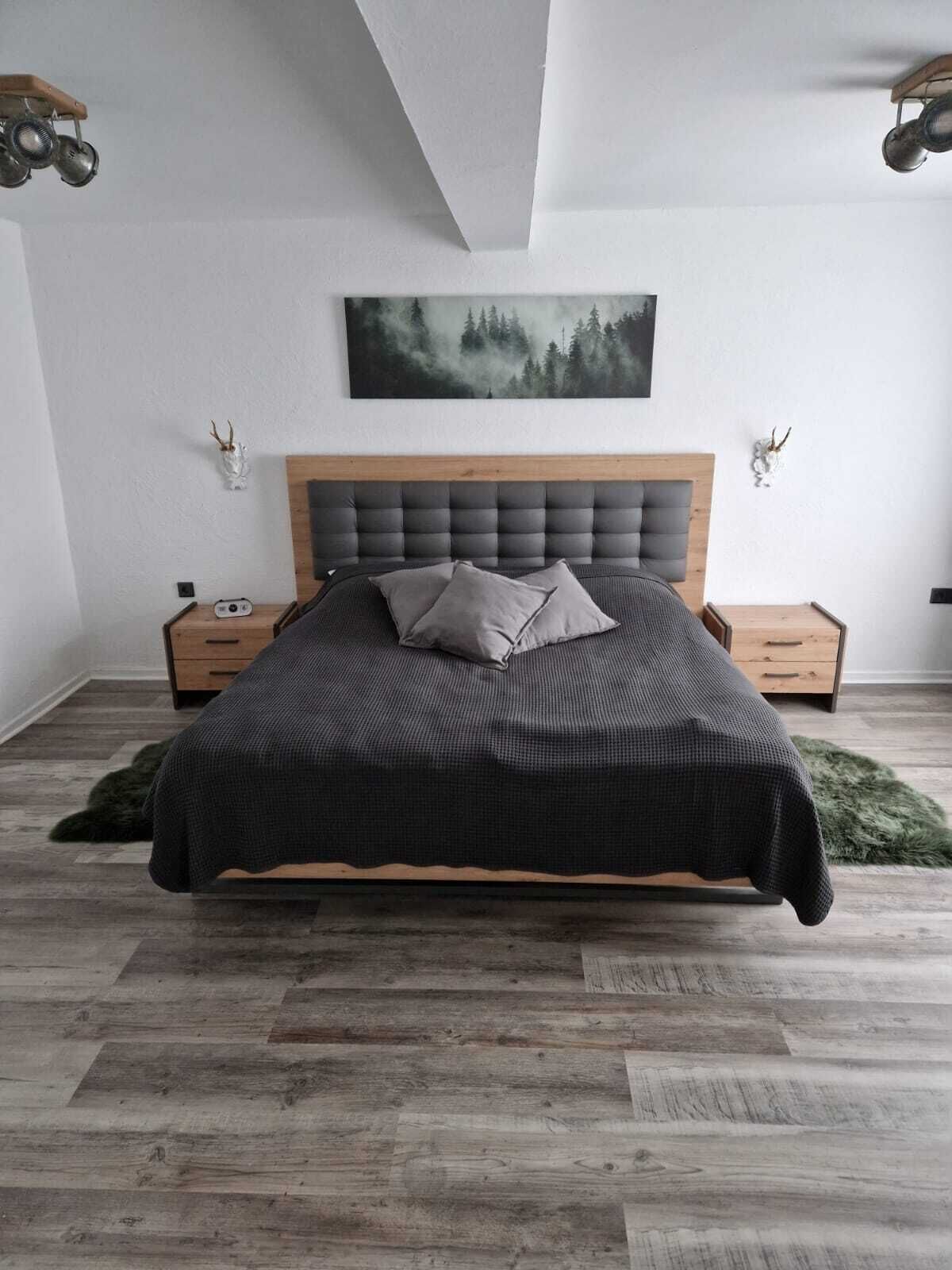 Un dormitorio moderno con cabecera de madera, una cama grande con colcha texturizada oscura, dos mesitas de noche y un cuadro con motivos forestales en la pared.