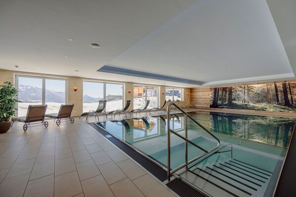 An indoor swimming pool with clear views of mountains through large windows, a wooden wall mural, and a potted plant. - Hotel Waldfrieden (Schladming)