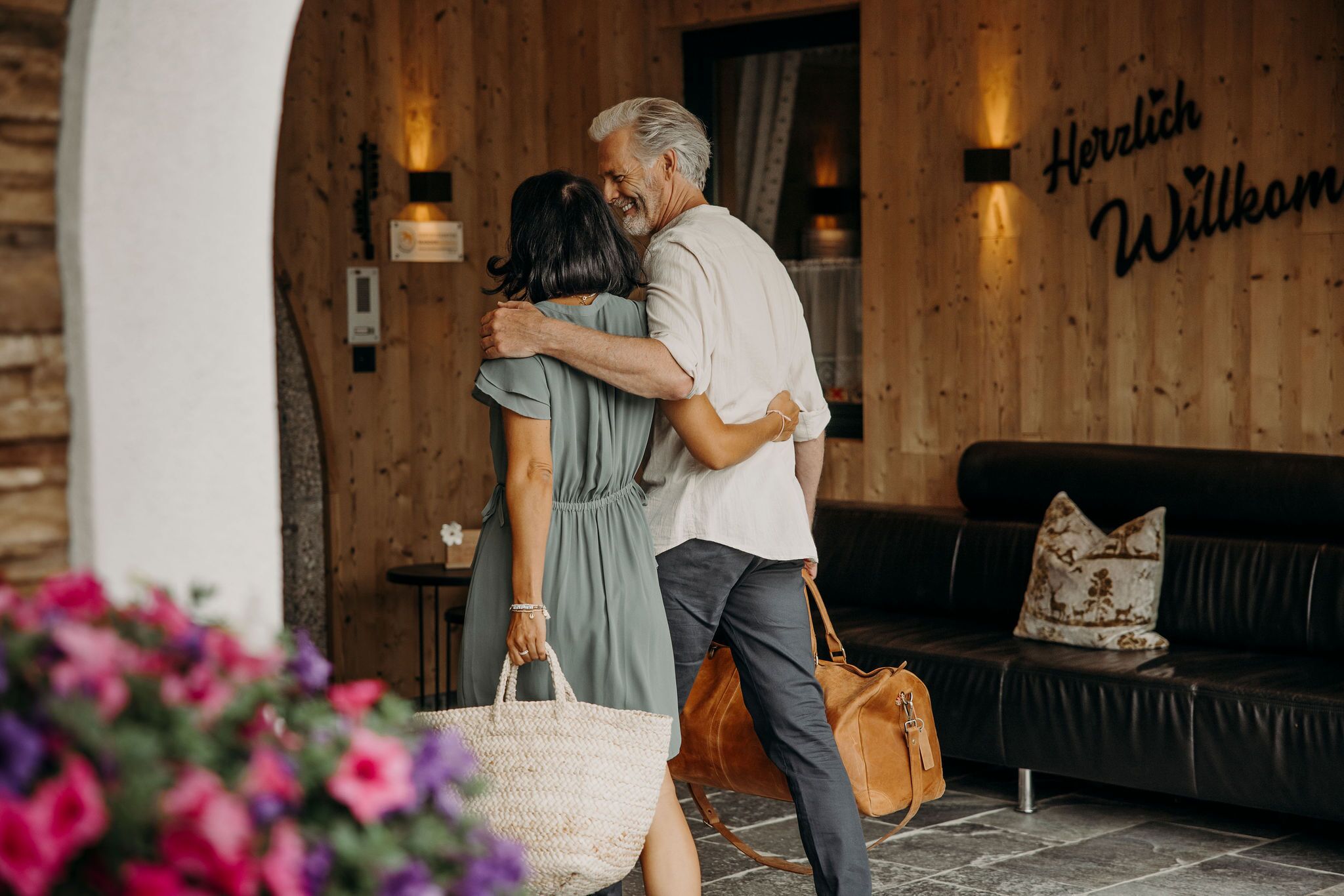 Un couple s’embrasse dans une pièce chaleureuse, éclairée d’une lumière douce, avec des murs en bois, un canapé en cuir noir et une pancarte indiquant « Bienvenue ».