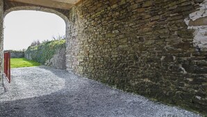 Un corredor de piedra con techo de madera, un camino de grava y un seto verde al fondo.