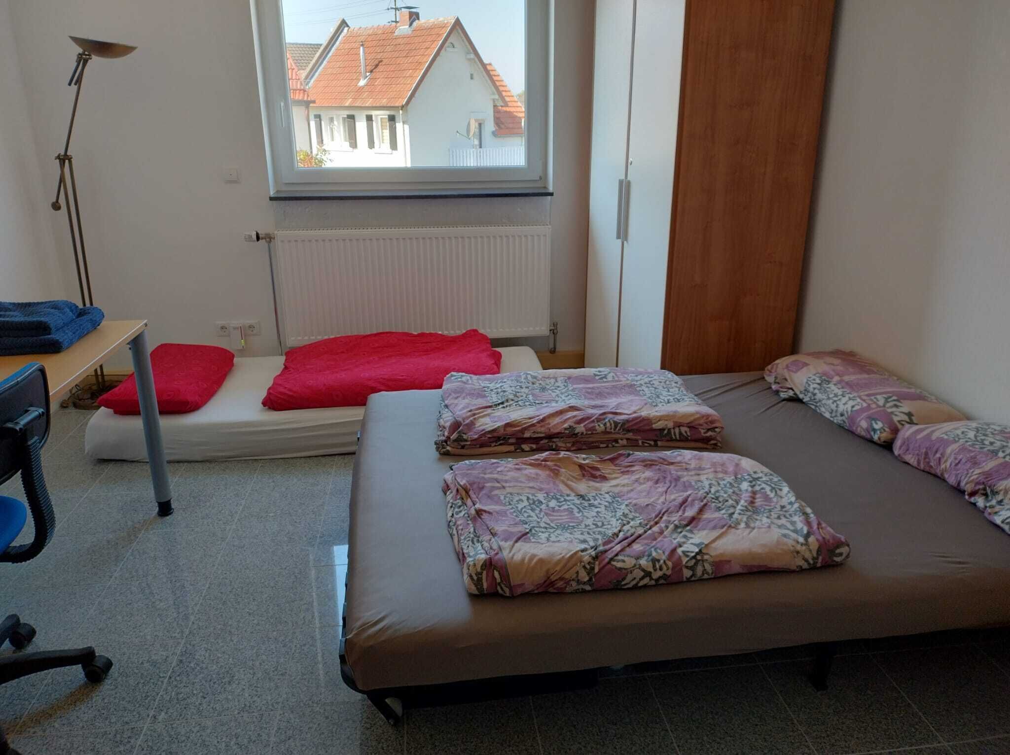 Um quarto pequeno e bem iluminado, com uma cama, uma escrivaninha e uma cadeira. Há uma janela que oferece vista para edifícios.