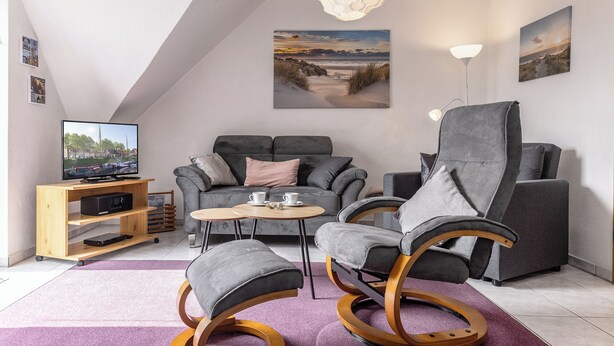A modern living room with a grey sofa, two grey armchairs, a wooden TV stand, and a purple rug.