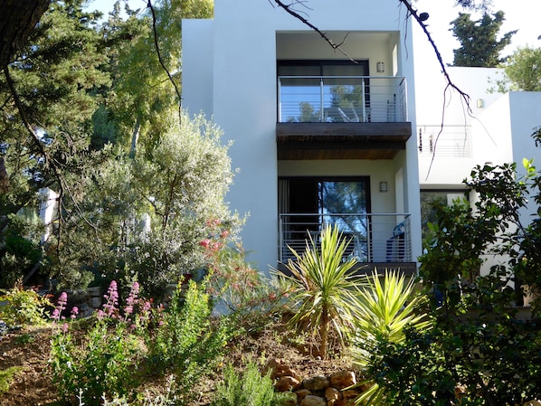 A modern two-story building with a balcony, surrounded by various plants and trees.