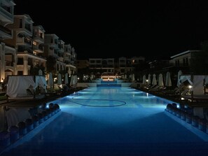 Ein Schwimmbecken bei Nacht mit Liegestühlen und Sonnenschirmen an beiden Seiten.