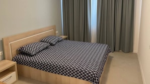 A neatly made bed with a geometric patterned bedspread, a wooden headboard, a bedside table with a drawer, and a wall-mounted air conditioner. - Sozopol Green life Apartment (Созопол)