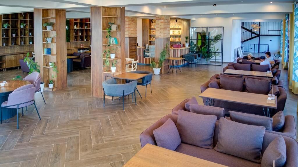 A spacious indoor area with wooden flooring, multiple seating arrangements including sofas and tables, and built-in wooden shelving units with plants.
