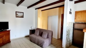 A living room with a sofa, a television on a wooden cabinet, a framed picture on the wall, and a kitchen area with a refrigerator. - Seaside in Roscoff, studio for 2 people, sea view, close to shops, beach (Roscoff)