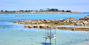 Eine felsige Küstenlinie mit klarem, blauem Wasser, einem Rettungsschwimmerstand und Gebäuden in der Ferne.